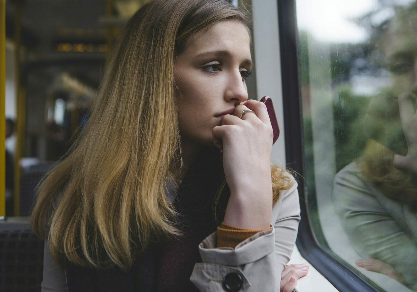 Young woman looking very upset as she sits on a train, looking out of the window. She is holding her phone in her hand up to her mouth.